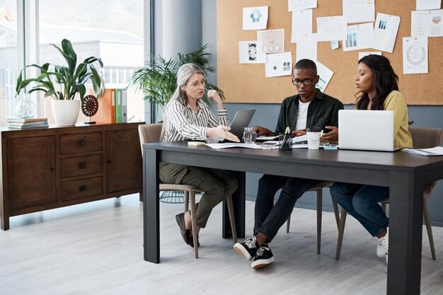 A group of diverse professionals sitting around a table in a modern office, engaged in a collaborative discussion. Digital devices like laptops and tablets are visible, suggesting the use of technology to enhance communication.